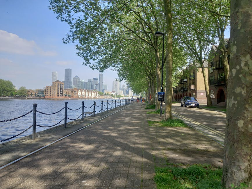 A wide paved pathway running alongside the River Thames in Canary Wharf, lined with mature trees providing dappled shade. On the right side, there are residential buildings with balconies made of brick and glass, adjacent to the pathway, and a black car is parked near the curb. Further along, a street lamp is visible among the trees, with a small mailbox attached to it. In the background across the river, the skyline features modern high-rise office buildings and skyscrapers characteristic of Canary Wharf, creating a contrast between the natural and urban environment. The scene is illuminated by natural daylight, with soft shadows cast by the trees, and the pathway appears to be part of a home relocation or moving process, as it is adjacent to a setup that could be used for transporting furniture or boxes during removals. Man with Van Canary Wharf's professional services are associated with such logistics, often involving careful packing, loading, and transport of possessions in this area.