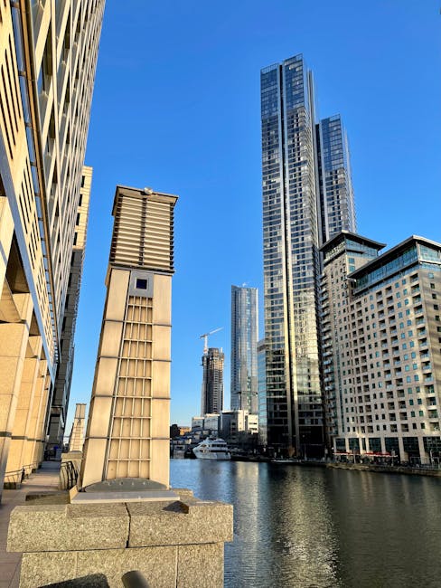 A view of a cityscape in Canary Wharf featuring modern high-rise buildings with glass facades, with a narrow waterway running between them. In the foreground, a concrete ledge supports a large vertical metal vent with wooden slats and a small rectangular control box attached. The scene is illuminated by bright daylight under a clear blue sky, illustrating an urban environment often associated with home relocation and furniture transport activities. The height and scale of the buildings highlight the need for professional removals services, such as those provided by Man with Van Canary Wharf, particularly during the loading and unloading process of moving household items or office equipment within this busy financial district.