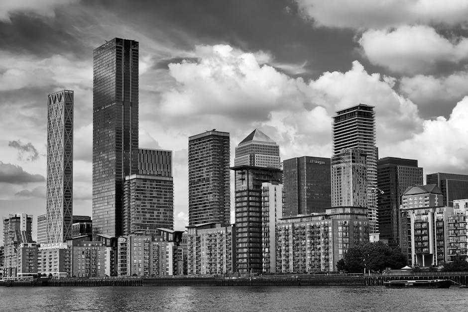 A black and white photograph of a cluster of modern high-rise buildings in a city skyline, viewed from across a body of water. The buildings vary in height, with some featuring distinctive architectural designs such as crisscross patterns and stepped facades. The lower section of the image shows a row of residential and commercial structures with balconies, situated along the waterfront. The sky overhead is filled with scattered clouds, creating a dynamic background. The scene is well-lit, highlighting the contrast between the glass and steel exteriors of the skyscrapers. This urban landscape may be part of a professional house removal or furniture transport operation, with a focus on the logistical aspect of loading and moving such large structures, which is occasionally handled by services like [COMPANY_NAME]. The image underscores the importance of efficient planning for relocation projects within a busy city environment, where careful handling of heavy furniture, packing materials, and loading equipment such as trolleys and straps may be involved in the process.