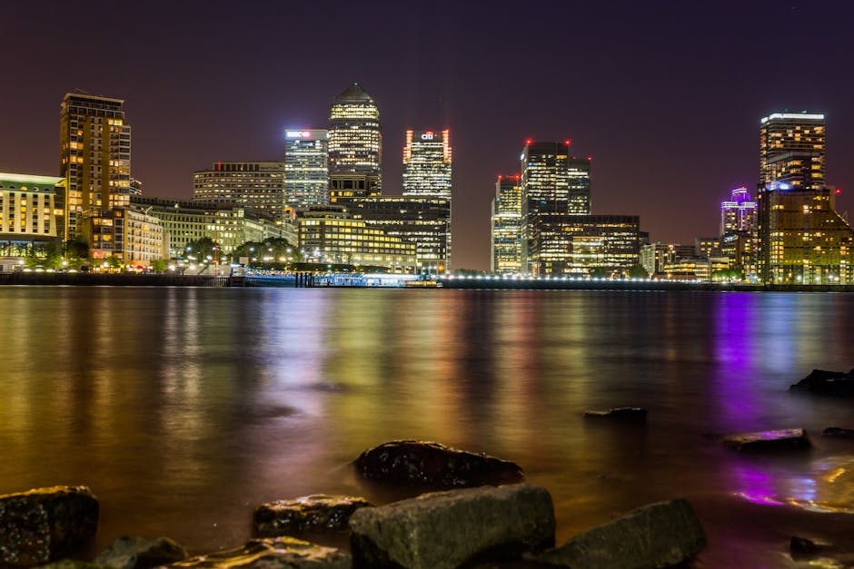 A nighttime cityscape of Canary Wharf featuring illuminated modern skyscrapers with reflective glass facades, situated along the riverbank. The buildings display various colours, including white, red, and purple lighting, creating a vibrant skyline. The calm river in the foreground reflects the lights from the urban area, with gentle ripples slightly distorting the reflections. Large rocks are visible along the water's edge, with some partially submerged. The scene is captured from a vantage point on the riverside promenade, with a few dark rocks in the foreground. The overall setting highlights the energetic and well-lit environment of Canary Wharf, relevant to home relocation and furniture transport services provided by Man with Van Canary Wharf during moving projects such as packing, loading, and transporting items to new addresses.