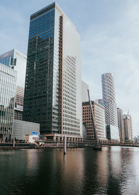 A black and white photograph of a cluster of modern high-rise buildings in a city skyline, viewed from across a body of water. The buildings vary in height, with some featuring distinctive architectural designs such as crisscross patterns and stepped facades. The lower section of the image shows a row of residential and commercial structures with balconies, situated along the waterfront. The sky overhead is filled with scattered clouds, creating a dynamic background. The scene is well-lit, highlighting the contrast between the glass and steel exteriors of the skyscrapers. This urban landscape may be part of a professional house removal or furniture transport operation, with a focus on the logistical aspect of loading and moving such large structures, which is occasionally handled by services like [COMPANY_NAME]. The image underscores the importance of efficient planning for relocation projects within a busy city environment, where careful handling of heavy furniture, packing materials, and loading equipment such as trolleys and straps may be involved in the process.