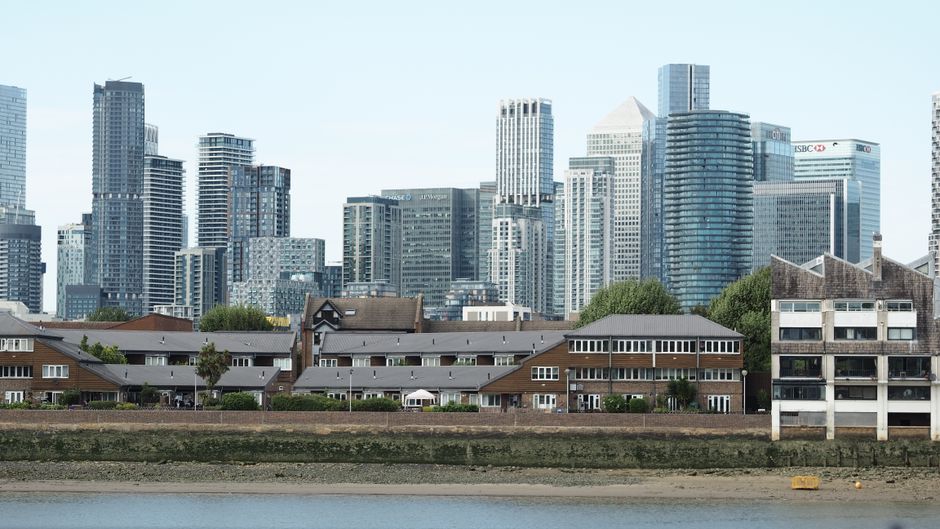 A view of a residential area with brown wooden houses and modern multi-storey apartment buildings beyond, situated along a riverbank in Canary Wharf. The wooden houses have grey roofs, windows, and sm