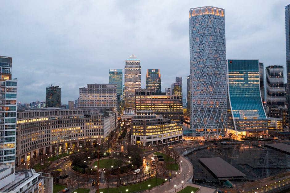 A view of a residential area with brown wooden houses and modern multi-storey apartment buildings beyond, situated along a riverbank in Canary Wharf. The wooden houses have grey roofs, windows, and sm
