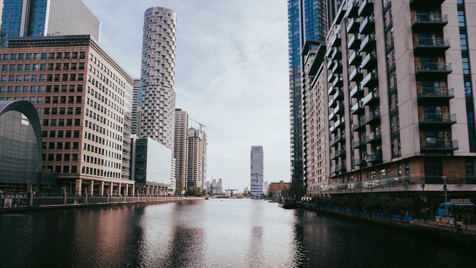 A cityscape view of Canary Wharf in London during dusk, featuring illuminated modern skyscrapers with glass facades and distinctive architectural designs, including the distinctive curved and lattice